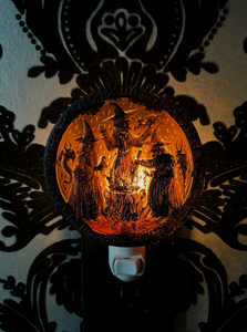 A circular Halloween night light with a carved witch and ghosts design, glowing with warm orange light from within, mounted on a white plug against a black and beige damask patterned wall.