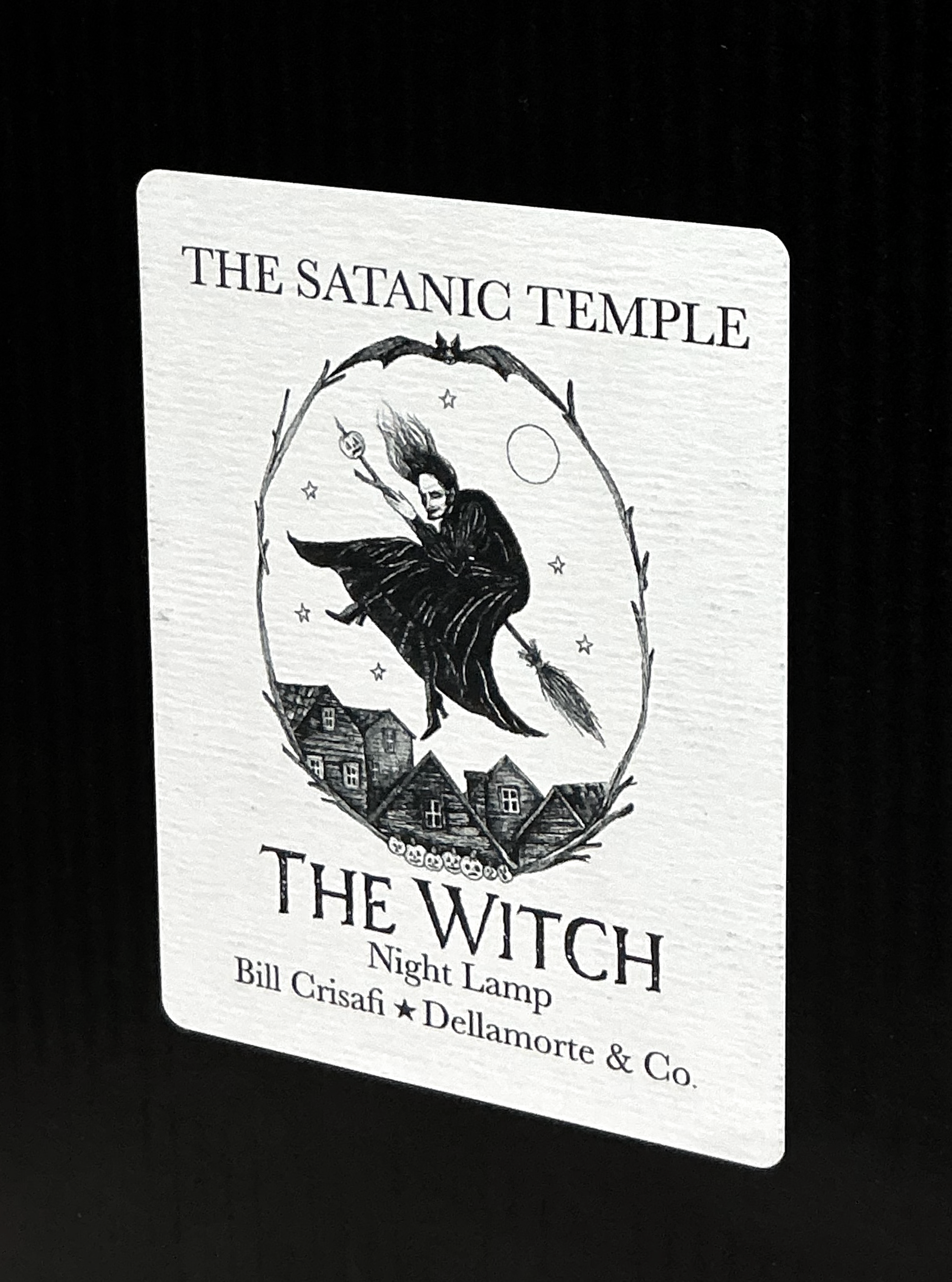 A white rectangular product card or label with black printed text reading 'THE SATANIC TEMPLE' and 'THE WITCH Night Lamp,' plus the names Bill Crisafi and Dellamorte & Co., shown against a black background.