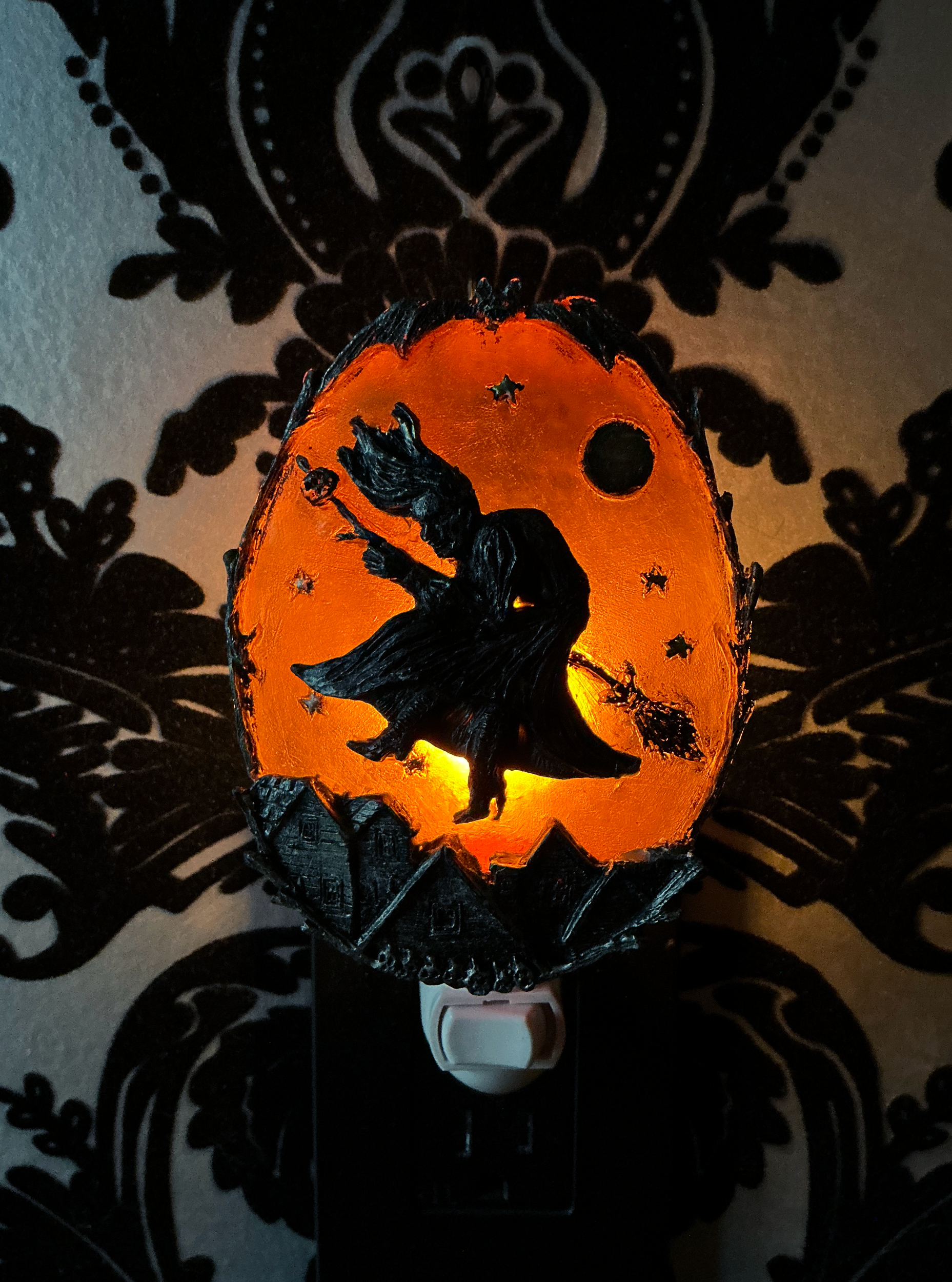 A Halloween night light shaped like a cracked egg, featuring a black silhouette of a witch riding a broomstick against a glowing orange background with a moon and stars. The night light is plugged into a wall outlet against a black and white damask wallpaper background.