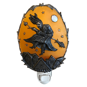 A Halloween-themed night light featuring an orange oval panel with a black metal relief of a witch riding a broomstick under a full moon, above small haunted houses, with a white switch base.