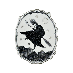 A silver-toned and white oval pendant or featuring a black illustration of a witch riding a broomstick over small houses under a full moon, with stars around the scene. Glows in the dark 