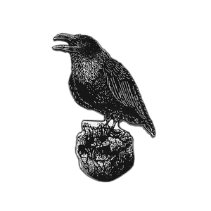 A black metal raven pin or charm with detailed feather texture, perched on a rock base, shown on a plain white background.