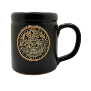 A glossy black ceramic mug featuring a circular gold-toned Norse-style emblem on the front with 3 witches, set against a plain white background.